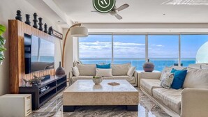 A modern living room with a large TV, comfortable sofas, a coffee table, and a view of the ocean.