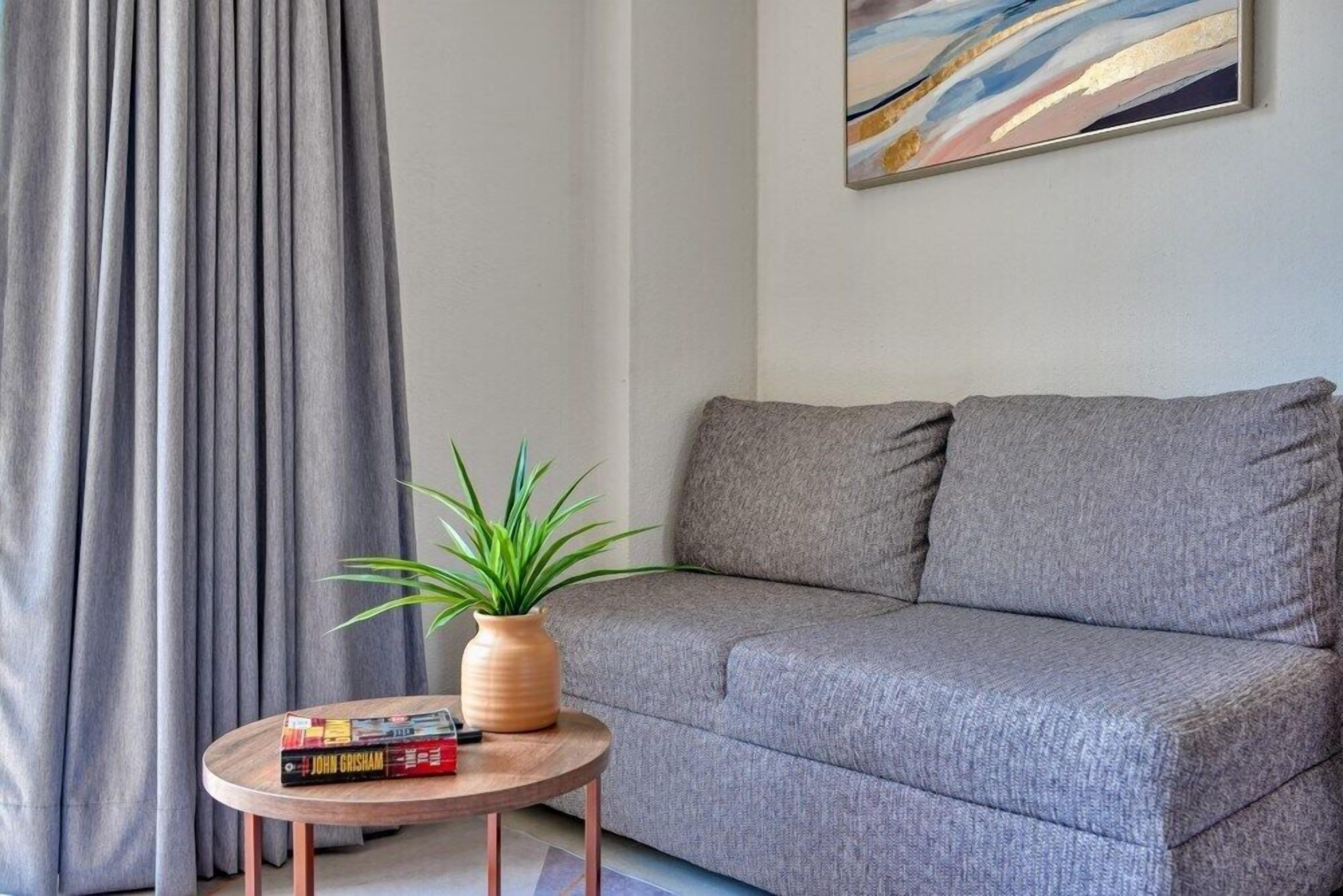 Un sofà gris, una taula rodona de fusta amb una planta i llibres, i un quadre a la paret.