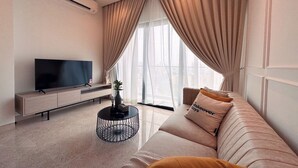 Een moderne woonkamer met een flatscreen televisie, een bank met kussens, een salontafel en een groot raam met gordijnen.