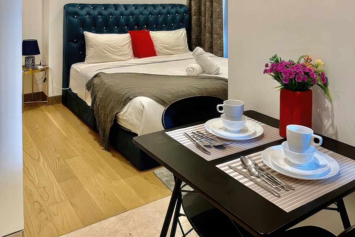 A modern hotel room with a tufted blue headboard, a bed with white and red pillows, a dining table with a vase of flowers, and a wall clock.