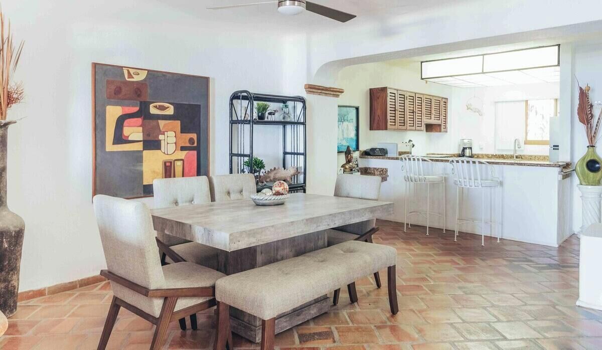 A modern kitchen with a dining area, featuring a large table, chairs, and a bench. There is a kitchen island with stools, a refrigerator, and a sink.