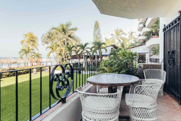 Un balcon avec une table ronde et des chaises en osier blanc, donnant sur un espace verdoyant agrémenté de palmiers et sur lequel on aperçoit un bâtiment au loin.