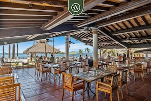 Un restaurant en plein air avec des tables et des chaises en bois, un toit de chaume et une vue sur la mer.