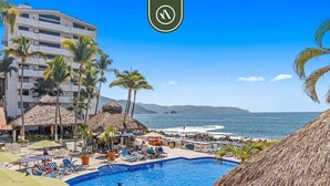 Zona de piscina en un resort con vista despejada al océano y montañas al fondo, rodeada de palmeras y cabañas con techo de paja.