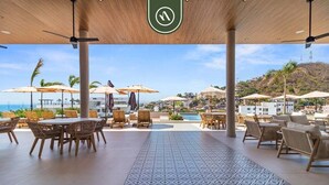 Een overdekt terras met tafels en stoelen, parasols en uitzicht op een zwembad en gebouwen op de achtergrond.