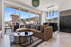 Un salon moderne avec un canapé d’angle, une table basse ronde et une vue sur un clocher, visible à travers de grandes fenêtres.