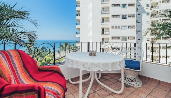 Uma varanda com uma mesa e cadeira brancas, uma manta colorida e vista para o mar e um edifício branco.