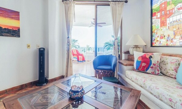 A living room with a glass coffee table, a sofa with floral cushions, a blue armchair, and a view through a glass door to an outdoor area with a red chair and a table. - 604 Vista del Sol | 2BR/2BA Ocean View, Beachfront (Puerto Vallarta)