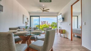 A modern living room with a glass dining table, grey upholstered chairs, a ceiling fan, and a view of buildings and greenery outside. - Brand New 2BR Apt - Infinity Pool - Versalles (Puerto Vallarta)