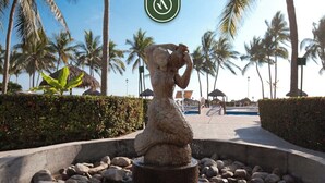 A stone statue in a water feature surrounded by rocks, palm trees, and a pathway. - Playa Royale Tower 1 1133 | 1BR Nuevo Vallarta | Beach, pool & WiFi (Nuevo Vallarta)