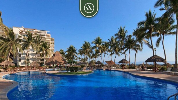 A resort pool with palm trees, a thatched-roof structure, and a hotel building in the background. - Playa Royale Tower 1 1133 | 1BR Nuevo Vallarta | Beach, pool & WiFi (Nuevo Vallarta)
