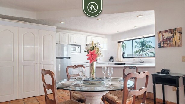 Un comedor con una mesa de vidrio rodeada de sillas de mimbre, armarios blancos y una cocina al fondo.