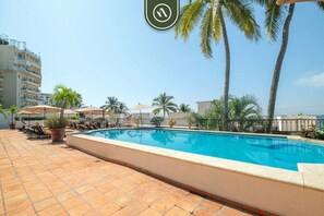 Ein Swimmingpool mit Liegestühlen und Sonnenschirmen, umgeben von Palmen und einem Gebäude im Hintergrund.