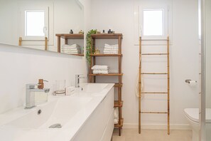 Un baño con un lavamanos blanco, espejo e inodoro. Hay estantes de madera con toallas y artículos de aseo. Una escalera de bambú está junto al inodoro.