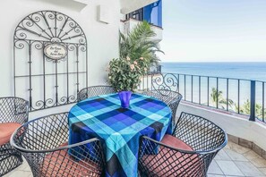 Een balkon met een geruit tafelkleed, metalen stoelen, een sierhek van metaal en uitzicht op zee.