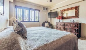 Een slaapkamer met een groot bed, een kaptafel, een aan de muur bevestigde televisie en een raam met tralies.