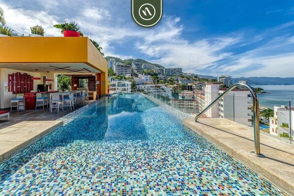 A swimming pool with mosaic tiles, a bar area with chairs, and a view of the city and sea.