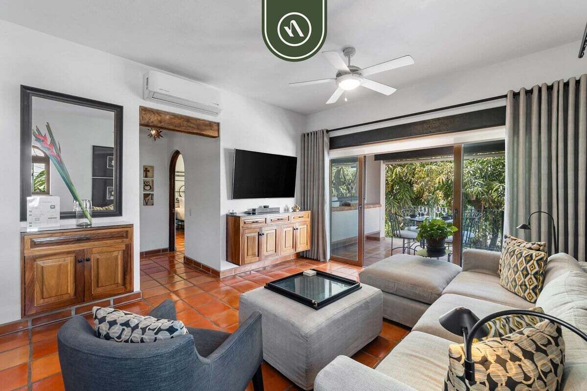 Una sala moderna con sofá, sillón y mesa de centro. Se observa un televisor, un mueble de madera y un ventilador de techo.