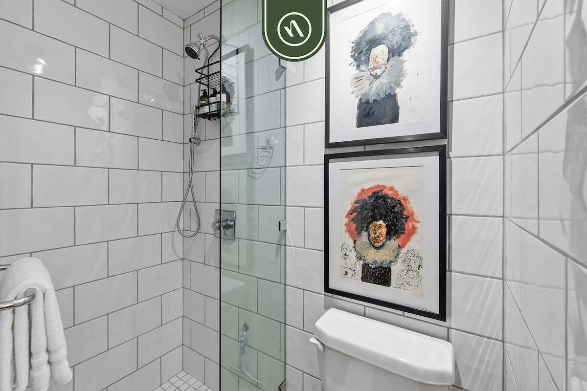 Een moderne badkamer met witte tegels, een glazen douchecabine, een toilet en twee ingelijste kunstwerken aan de muur.