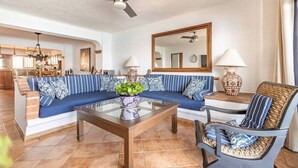 Un espacio de sala amplio con un conjunto de sofá, una mesa de centro y una silla. Se ve un ventilador de techo, y hay una cocina en el fondo.
