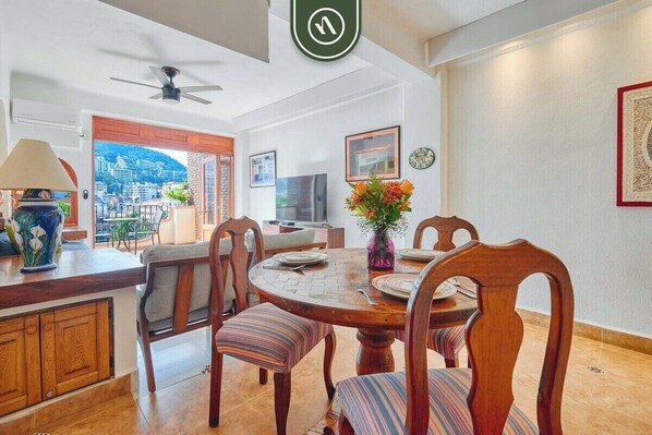 A dining area with a round wooden table, striped cushioned chairs, a vase with flowers, and a city view through an open door.