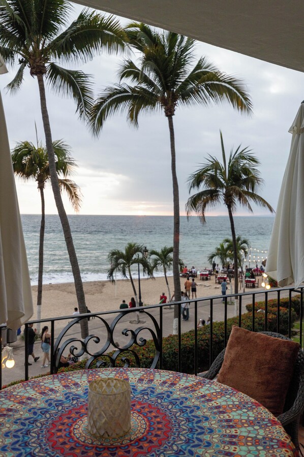 Ein Balkon mit einem gemusterten Tisch, einem braunen Kissen und Blick auf den Strand mit Palmen und dem Meer.