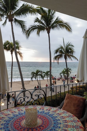 Ein Balkon mit einem gemusterten Tisch, einem braunen Kissen und Blick auf den Strand mit Palmen und dem Meer.