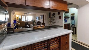 Una cocina con armarios de madera, encimera de mármol y un comedor al fondo.