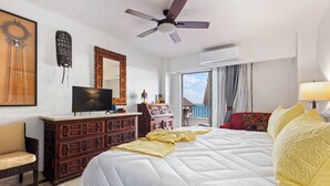 Habitación de hotel con cama, televisor, cómoda, silla, ventilador de techo y ventana con vistas.