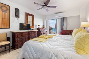 Ein Hotelzimmer mit Bett, Fernseher, Schminktisch, Stuhl, Deckenventilator und einem Fenster mit Blick.