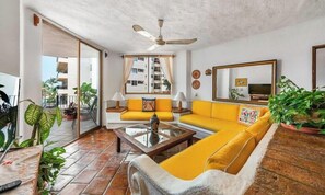 A living room with a yellow sofa, a glass coffee table, a ceiling fan, and a large mirror.
