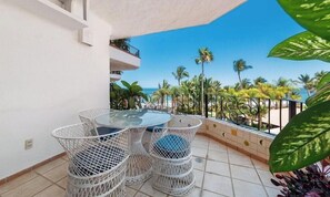 A balcony with a glass table and wicker chairs, overlooking a beach with palm trees and the ocean.