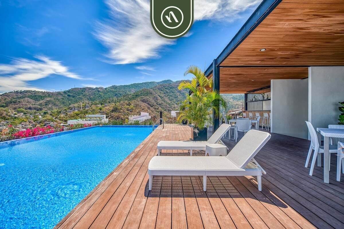 Uma área de piscina moderna com deck de madeira, cadeiras de lounge brancas e vista para uma paisagem montanhosa.