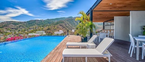 A modern pool area with wooden decking, white lounge chairs, and a view of a hilly landscape.