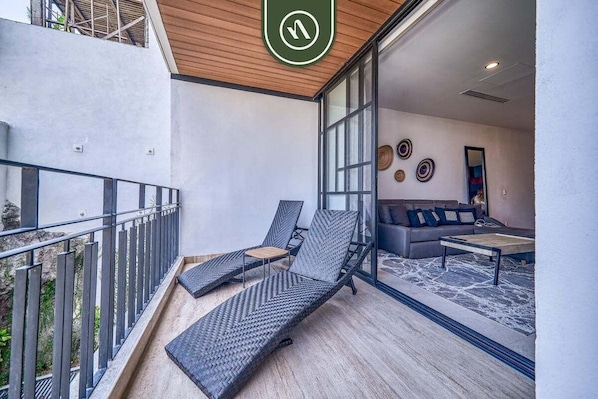 A balcony with two lounge chairs, a sofa, and a coffee table inside a modern room with large windows and a wooden ceiling.
