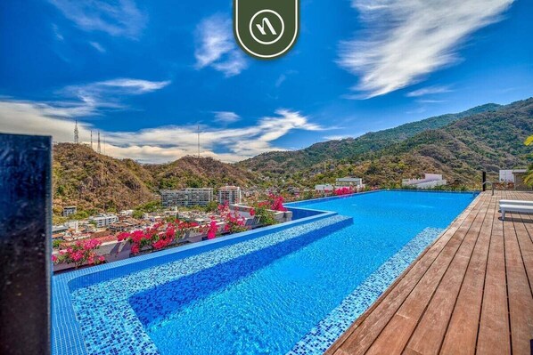 A swimming pool with a wooden deck, surrounded by hills and buildings.