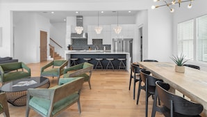 A modern kitchen with white cabinets, a central island, and bar stools. There is a dining area with a wooden table and chairs. - Walk to Beach + Pool, Hot Tub & Game Room | 30A Ohana Grande by AvantStay (Santa Rosa Beach)