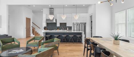 A modern kitchen with white cabinets, a central island, and bar stools. There is a dining area with a wooden table and chairs.