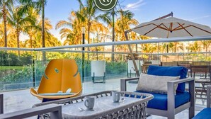 A modern outdoor seating area with a glass balustrade, a yellow chair, a blue sofa, and a white umbrella.