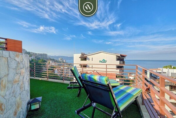 A rooftop terrace with a view of the sea, a lounge area with striped cushions, and a building with a logo.