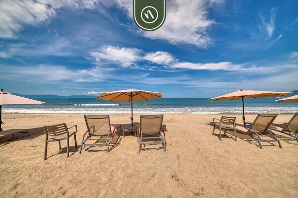 A beach with sun loungers and umbrellas, a clear sky, and the ocean in the background.