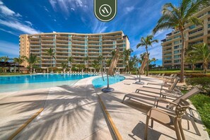 A hotel with a large swimming pool, sun loungers, and palm trees.