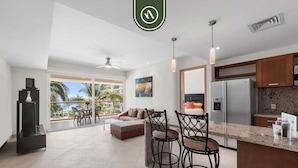 A modern kitchen with a refrigerator, a dining area with bar stools, and a large window offering a view of palm trees.
