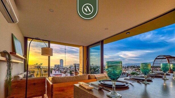 Ett modernt vardagsrum med ett stort fönster, en soffa, ett matbord dukat med glas och utsikt över en stadsbild i solnedgången.