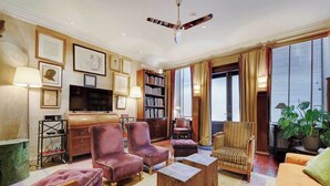 Una sala de estar bien iluminada con ventilador de techo, asientos cómodos, una estantería y un amplio ventanal con cortinas.