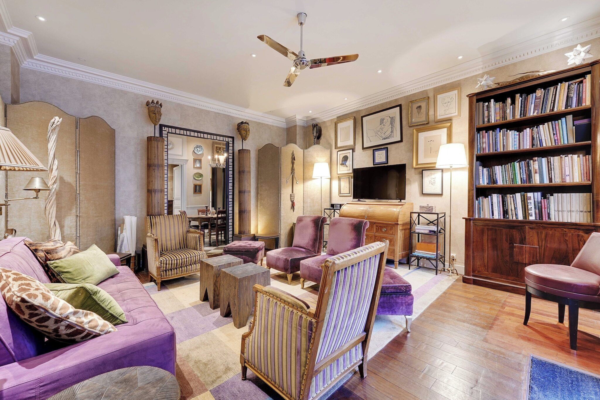 Een woonkamer met een paarse bank, fauteuils, een houten salontafel, een boekenkast en een televisie.