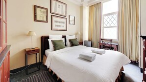 Habitación de hotel con una cama grande, dos mesitas de noche, un escritorio y una silla. Se observan cuadros enmarcados en la pared y una ventana con cortinas.