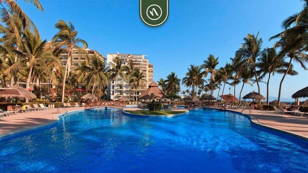 Ein großer Pool, umgeben von Palmen und Liegestühlen, mit einem Hotel im Hintergrund.