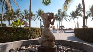 Eine Statue einer Frau in tropischer Umgebung, mit Palmen und einem Pool im Hintergrund.