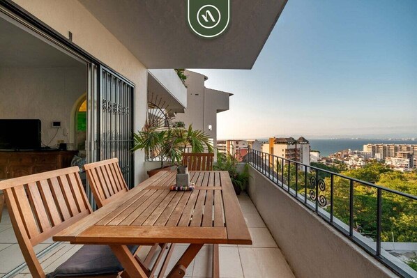 A balcony with a wooden table and chairs, a TV, and a view of the city and sea.